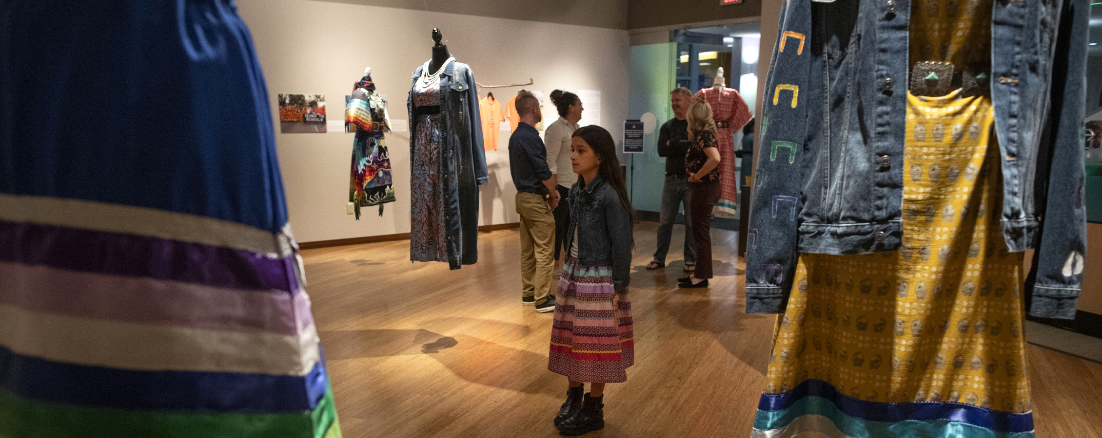 A child with long dark hair stands in Roberta's Art Gallery and looks at an exhibit of colorful clothing hanging from the ceiling which honors indigenous persons.